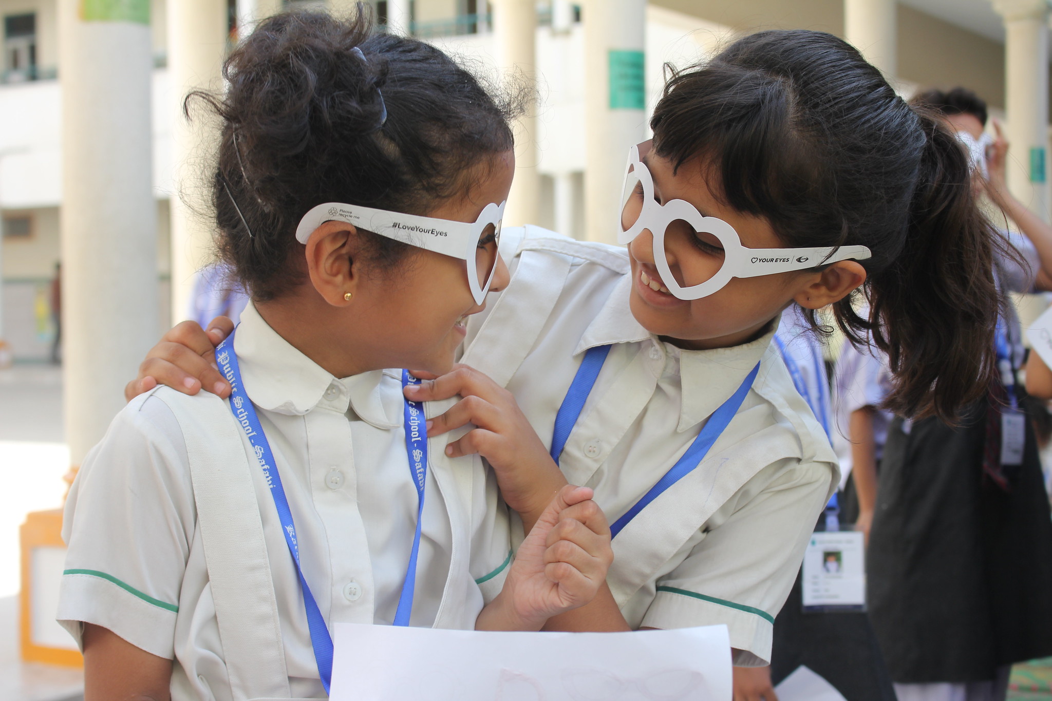 Two young girls wearing playful eye-shaped glasses smiling at each other during a school eye health event.